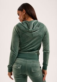 Felpa in velluto verde con cappuccio regolabile e orlo a costine, abbinata a pantaloni coordinati dotati di tasche posteriori e dettagli argentati.