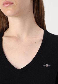 Črn pleten pulover z V-izrezom in rebrastim robom. Ima majhen vezen logotip na prsih. Gladka, mehka tekstura.