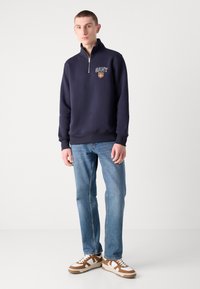 Hanorac cu fermoar de marine, cu logo brodat, asortat cu blugi de nuanță albastru deschis și adidași maro și alb. Design simplu și casual.