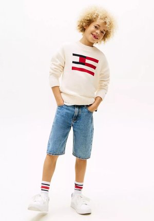 Maglione bianco lavorato a maglia con design a bandiera rossa e nera, pantaloni corti in denim blu, sneakers bianche e calze bianche con righe rosse. Capelli ricci.