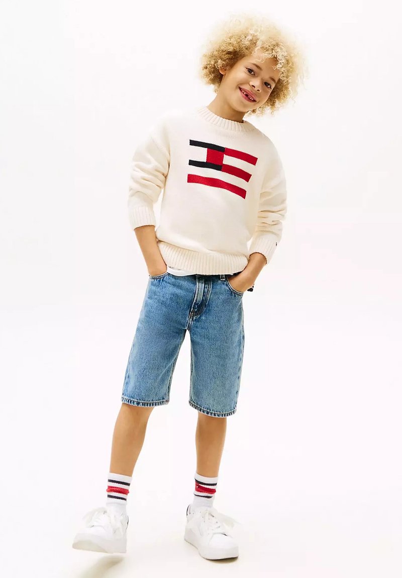 Maglione bianco lavorato a maglia con design a bandiera rossa e nera, pantaloni corti in denim blu, sneakers bianche e calze bianche con righe rosse. Capelli ricci.