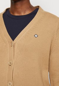 Beige strikket cardigan med V-hals, fire knapper og et lille logomærke på brystet. Brugt over en marineblå T-shirt.