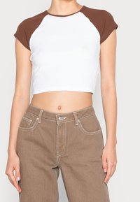 En person som bär en vit kortärmad t-shirt med bruna ärmar och högmidjade bruna byxor som står framför en neutral bakgrund.