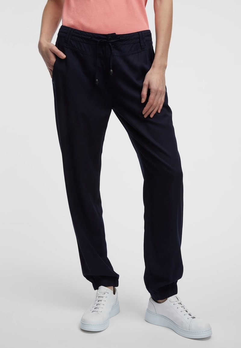 Femme portant un pantalon de jogging noir avec des chevilles élastiques, un haut corail et des baskets blanches, debout avec une main dans la poche.