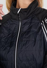 Gros plan d'une personne portant une veste matelassée noire à col montant et fermeture éclair à rayures blanches, ajustant le col avec la main gauche.