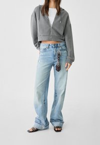 Grijze cropped hoodie met rits, gecombineerd met lichtblauwe distressed wijde jeans. Zwarte sandalen met vierkante neus en kettingaccenten.
