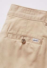 Pantalones Levi's beige con un bolsillo trasero con un botón marrón, detalles de costura y una etiqueta blanca con el nombre de la marca.