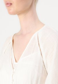 Blouse légère blanche présentant un décolleté en V superposé et un délicat feston en dentelle le long de l'épaule. Texture douce avec des boutons à l'avant.