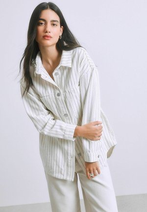 Jonge vrouw met lang donker haar, gekleed in een wit gestreept overhemd met knopen en witte broek, poserend tegen een effen lichte achtergrond.