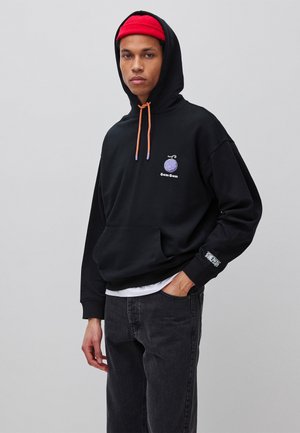 Junger Mann trägt schwarzen Hoodie mit lila Logo und orangen Kordeln, rote Mütze und schwarze Jeans, steht mit einer Hand in der Hosentasche.