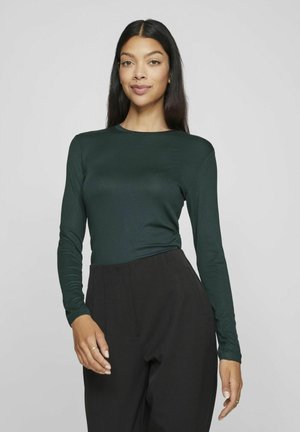 Blusa de manga larga de color verde oscuro fabricada en tejido suave, con escote redondo y silueta ajustada, combinada con pantalones negros de cintura alta.