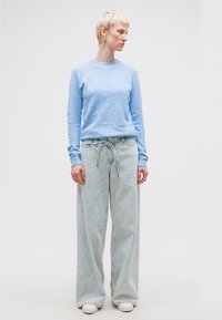 Άτομο με κοντά ξανθά μαλλιά που φοράει ανοιχτό μπλε πουλόβερ, φαρδιά τζιν με λάστιχο στη μέση και λευκά αθλητικά παπούτσια, στέκεται μπροστά σε απλό φόντο.