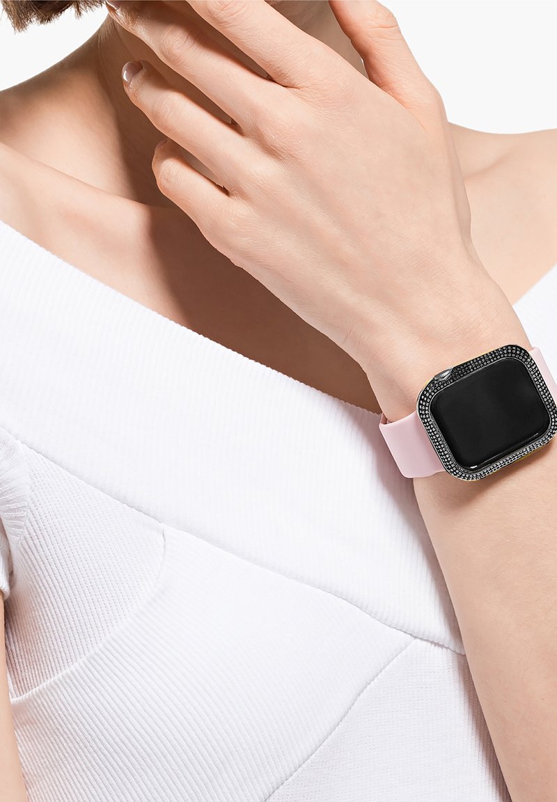 Femme portant un haut blanc à épaules dénudées, main près du visage, avec une montre connectée noire dotée d'un bracelet rose et d'une lunette ornée de cristaux au poignet.