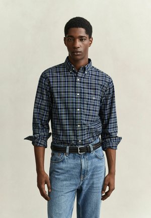 Jeune homme portant une chemise à carreaux bleue à manches retroussées et un jean bleu clair sur fond neutre.