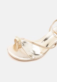 Χρυσό μεταλλικό σανδάλι ανοιχτού τύπου με λεπτά λουράκια και δαχτυλίδι για το δάχτυλο διακοσμημένο με μικρές στρας.