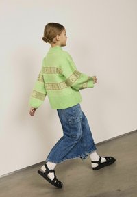 Green knitted sweater with ribbed hem, featuring light brown stripes with text. Paired with wide blue denim pants and black sandals.