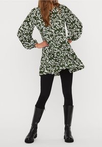 Robe verte à motifs floraux avec manches longues bouffantes et ourlet volanté, portée avec des leggings noirs et des bottes noires montantes. Présente des motifs floraux blancs.