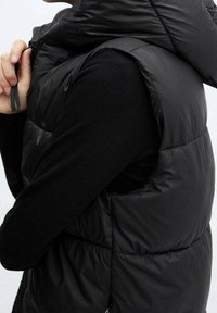 Veste matelassée noire avec col montant, fermeture zippée à l'avant et design sans manches. Présente une texture lisse et des coutures horizontales.