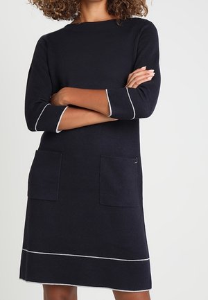 Femme portant une robe noire à manches longues avec deux poches devant et un liseré blanc aux manches et à l'ourlet, bras croisés.