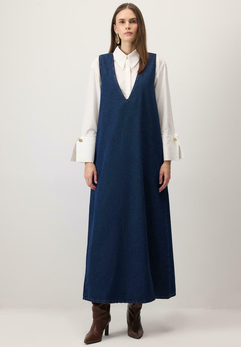 Tumši zila džinsa maksi kleita ar dziļu V veida izgriezumu un platām padusēm, pārģērbta pāri baltai kreklam ar zvana formas piedurknēm un pogu akcentiem.