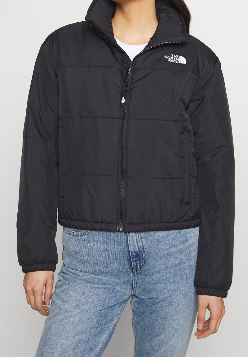 Personne portant une doudoune noire avec un col montant et un logo sur la poitrine, associée à un jean en denim bleu clair.