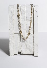 Gold and silver-tone metal chain necklace with alternating round and oval links, displayed on a textured white pedestal.