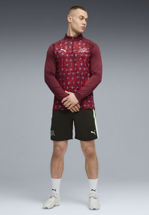 Nuori mies, jolla on yllään viininpunainen, kuvioitu vetoketjutakki, mustat Puma-shortsit, valkoiset Puma-sukat ja valkoiset nappikengät. Hän seisoo käsivarret ristissä ja katsoo vasemmalle.