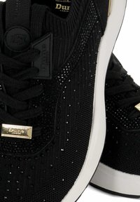 Baskets noires en matériau tricoté texturé avec des accents scintillants, des détails en or et une semelle blanche. Les lacets sont noirs avec un patch logo.