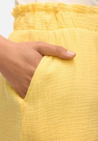 Żółta, fakturowana tkanina z warstwowym pasem w falbany, z boczną kieszenią i miękką, pomarszczoną powierzchnią. Zbliżenie na rękę w kieszeni.