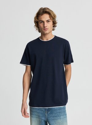 Młody mężczyzna z kręconymi włosami, ubrany w granatowy t-shirt z białym wykończeniem i jasnoniebieskie dżinsy, stoi na jednolitym, szarym tle.