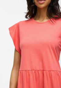 Robe corail à manches courtes et flottantes, décolleté rond et taille froncée pour un ajustement ample. Tissu en coton doux, texture lisse.