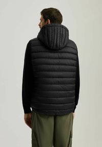 Czarna ocieplana kamizelka z kapturem, z pikowaną fakturą i poziomymi pasami, noszona na długim rękawie i zielonych spodniach cargo.