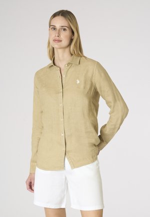 Donna che indossa una camicia beige a maniche lunghe con bottoni e pantaloncini bianchi, in piedi con una mano dietro la schiena su uno sfondo semplice.