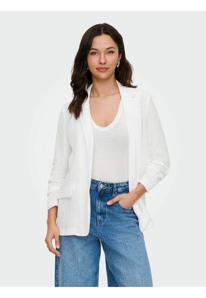 Frau mit langen dunklen Haaren, die einen weißen Blazer, ein weißes Top, blaue Jeans und goldene Creolen trägt und mit einer Hand in der Tasche posiert.