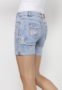 Shorts de mezclilla azul claro con estampado floral en tonos de azul y beige; dobladillo enrollado; dos bolsillos traseros con remaches plateados.