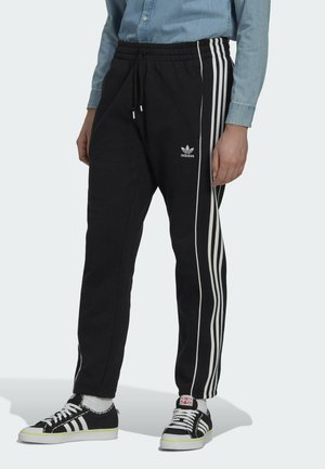 Pantalones deportivos negros con franjas laterales blancas, cintura elástica y cordón. Combinados con zapatillas negras y calcetines blancos.