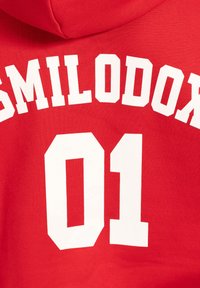Červená mikina s kapucí, která na zádech má velký bílý nápis "SMILODON" a číslo "01". Měkká, texturovaná tkanina.