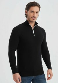 Pull en maille noire avec col montant, doté d'une fermeture éclair argentée, coupe ajustée et manches longues, associé à un jean bleu.
