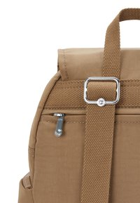 Hnědý látkový batoh s nastavitelným popruhem, stříbrnou přezkou a malou zipovou kapsou viditelnou na bílém pozadí.