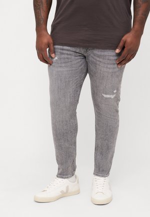 Homme portant un jean skinny gris délavé, des baskets blanches et une chemise marron foncé, debout devant un fond blanc.