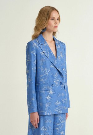 Donna che indossa un blazer doppiopetto floreale blu e pantaloni abbinati, in piedi contro uno sfondo chiaro e uniforme, vista di profilo.
