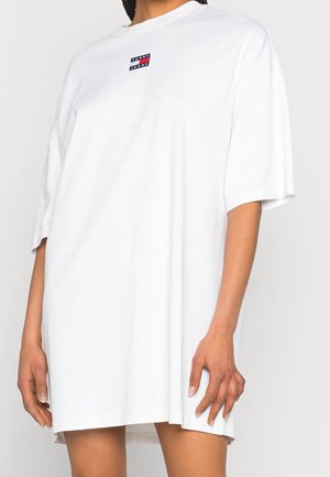 Vrouw draagt een oversized witte T-shirtjurk met een klein rechthoekig logo op de borst, staand met de armen ontspannen langs het lichaam.