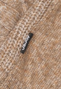 Pull en maille aux tons terreux de marron et de beige, avec un tissu texturé et une petite étiquette noire avec un texte blanc cousue sur le côté.