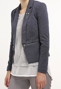 Mörkblå blazer med skräddarsydd passform, fickor med dragkedja och en mjuk textur, bärs över en ljusgrå topp med lageredd fåll.