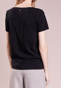 Black short-sleeve t-shirt with a rounded neckline, cotton material, featuring a small logo at the back. Worn with light gray pants.