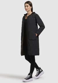 Cappotto lungo nero imbottito con un motivo a rombi trapuntato, dotato di cappuccio e due tasche frontali, indossato su un top e leggings neri.