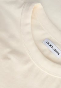 Κοντινή λήψη ενός βαμβακερού T-shirt σε κρεμ απόχρωση με ορατή ετικέτα της μάρκας Jack & Jones στο εσωτερικό του γιακά.