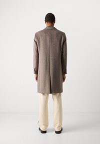 Abrigo marrón a cuadros con silueta estructurada, solapas en muesca y puños abotonados, combinado con pantalones color crema y zapatos negros.