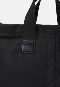Borsa tote nera realizzata in tessuto liscio con spalline nere a contrasto e un'etichetta logo texturizzata. Presenta un design minimalista con linee pulite.