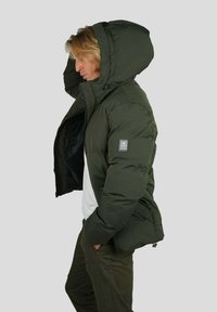 Personne portant une veste matelassée vert foncé avec la capuche levée, une chemise blanche et un pantalon vert assorti, se tenant de profil avec les mains dans les poches.